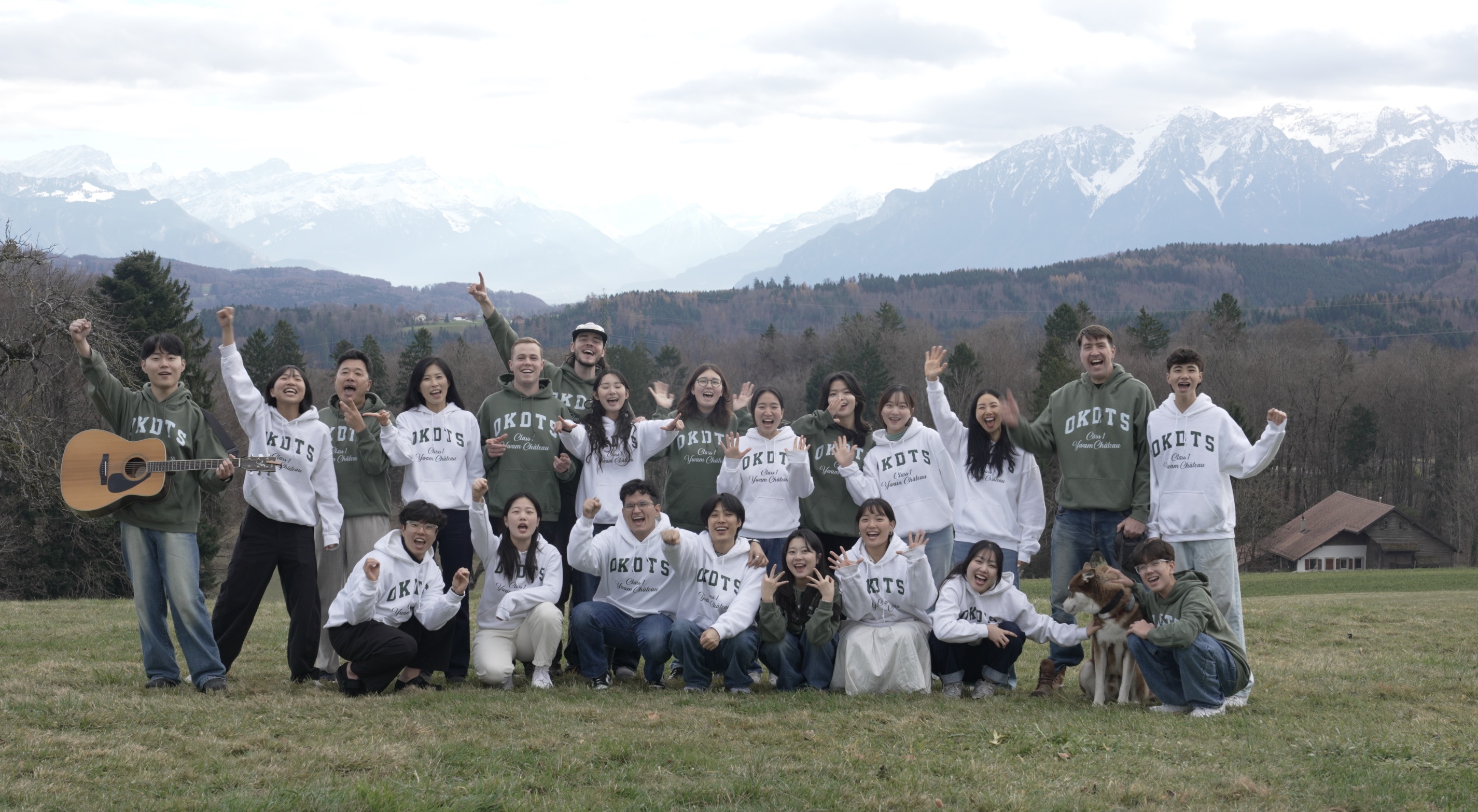 OneKorea DTS Class 1: students and staff in OKDTS hoodies celebrating together in an alpine meadow, Swiss mountains behind