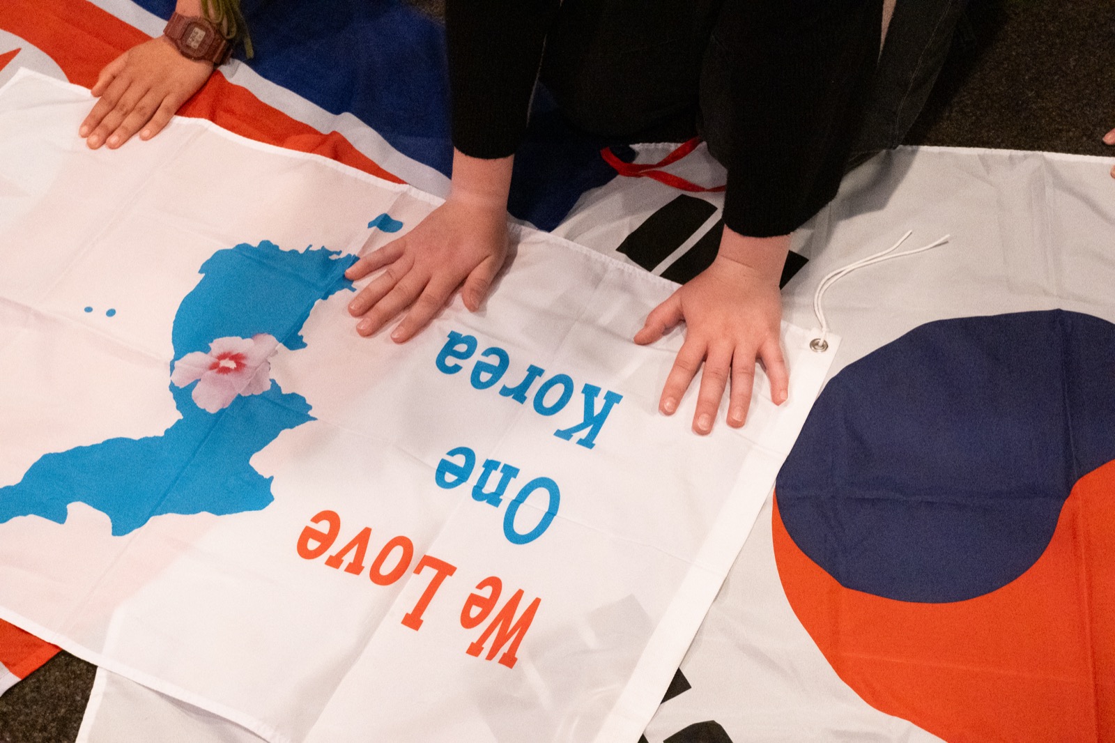 Hands pressed over a We Love One Korea banner on the floor, Korean peninsula map visible