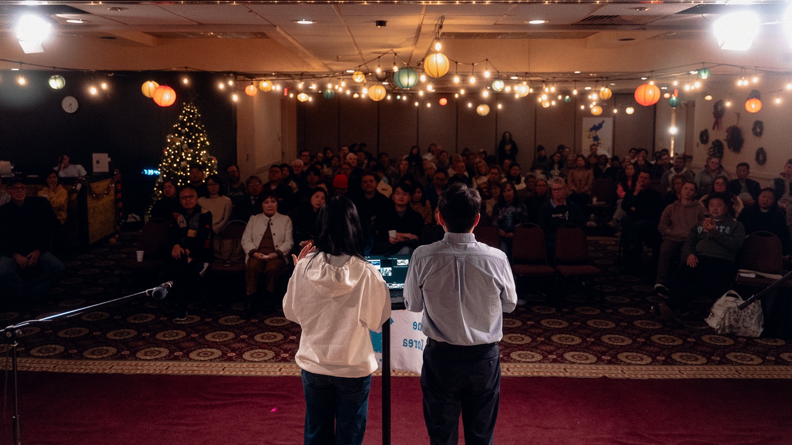 Two speakers facing a full audience at an OK Vision Night in Colorado Springs, warm string lights and lanterns overhead